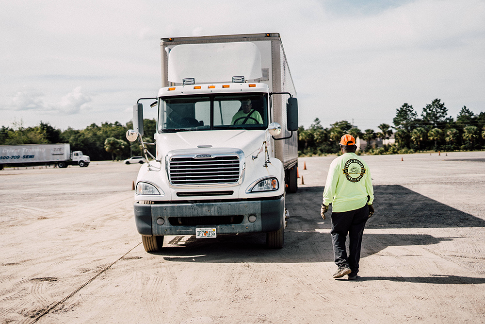 A man in a neon yellow sweatshirt and orange baseball cap walks up to a large white truck in a parking lot.