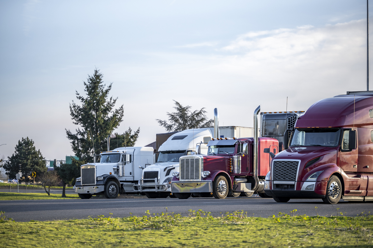 Different models of big rigs semi trucks tractors with car hauler and refrigerator and dry van semi trailers standing in row on the truck stop industrial parking lot for the rest at twilight time