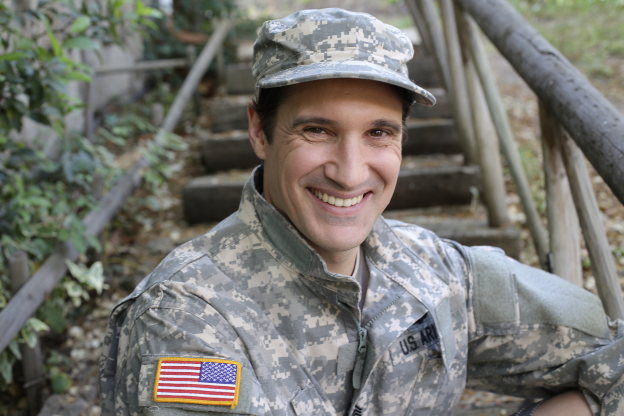 A caucasian army soldier in his 30s is  wearing a traditional USA army uniform with cap. He looks at camera and is outdoors.