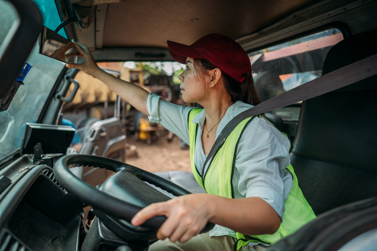 Personal perspective of mid 30s woman in casual clothing, cap, and reflective vest sitting in driver’s seat, adjust mirror and looking straight ahead, ready to begin road trip.