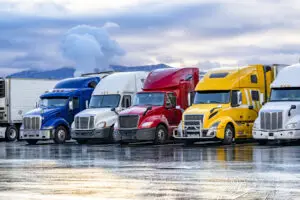 Different make big rigs semi trucks tractors with loaded semi trailers standing in the row on truck stop parking lot at early morning waiting for the route continuation time according to the log book