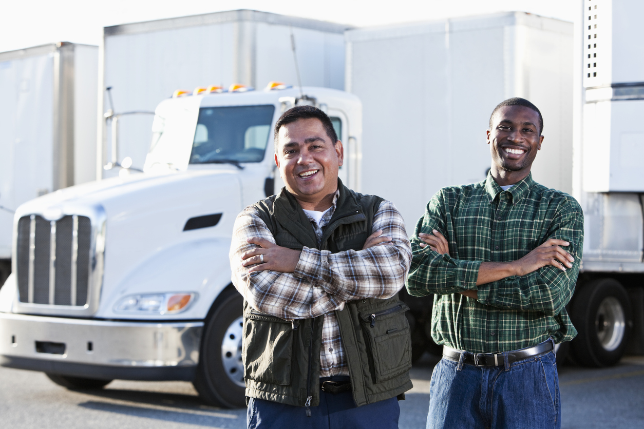 Hispanic and African American truck drivers standing in front of semi-trucks.