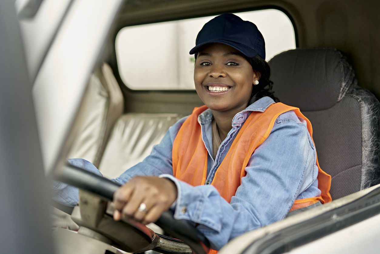 Three-quarter front view of mid 20s woman in denim shirt, cap, and reflective vest sitting in driver’s seat with hands on steering wheel and smiling at camera.
