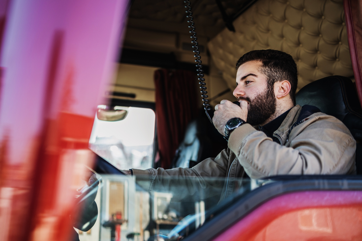 Young handsome bearded man driving his truck. He is using built in cabin CB radio to communicate with other drivers and control center.