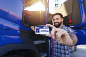 Portrait of middle aged bearded truck driver standing by his truck and showing his commercial driver license. Focus on CDL license. Truck driving school and job openings.