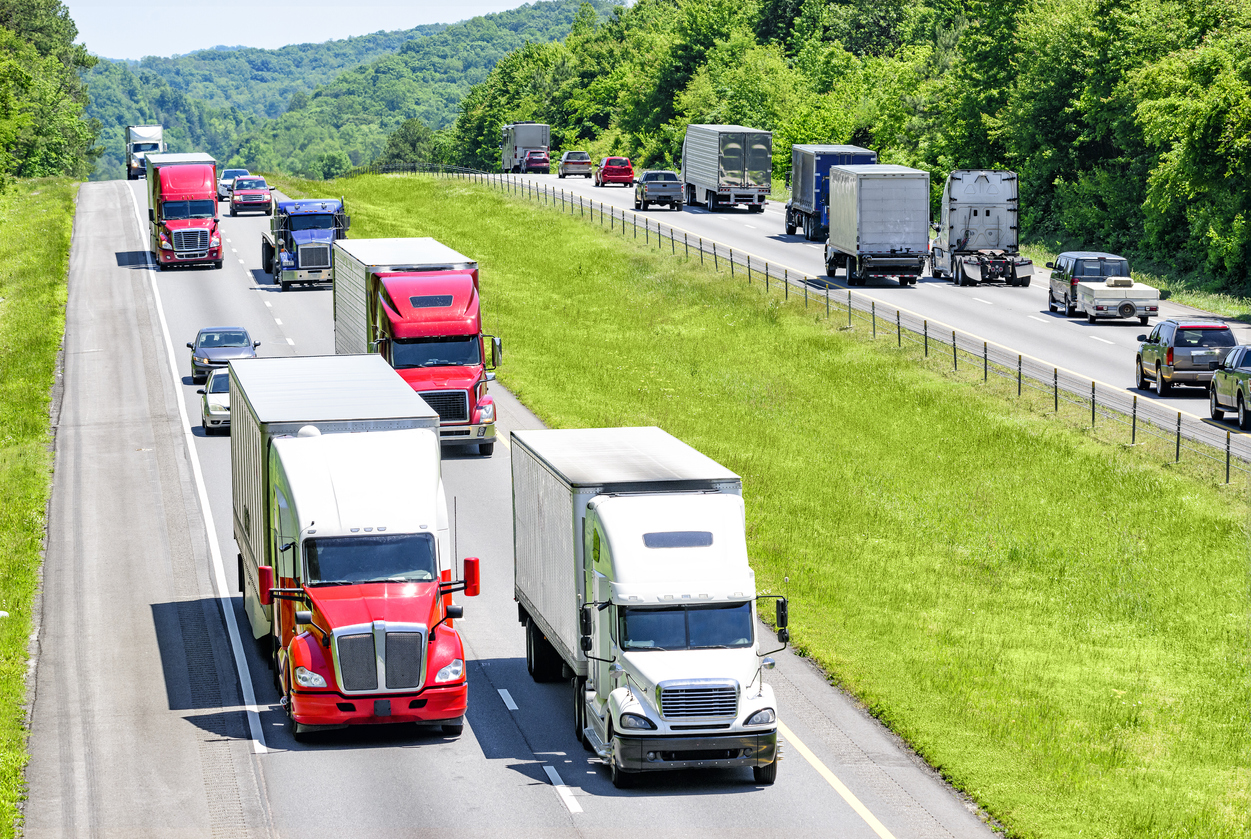 A heavy flow of 18-wheelers peppered with cars and SUVs roll down a Tennessee interstate highway. Heat rising from the pavement gives a shimmering effect to vehicles and background in the distance.
