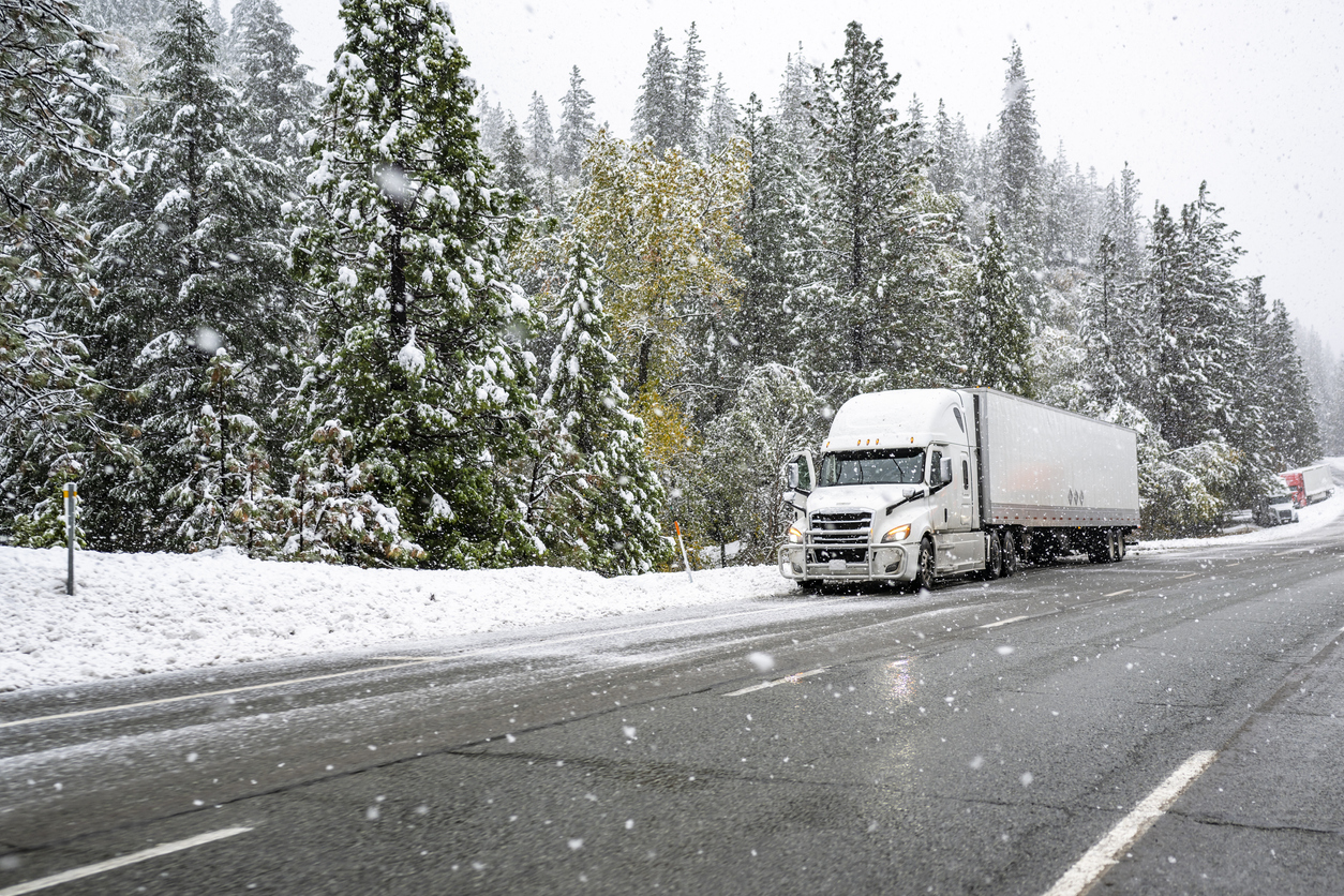 White big rig industrial semi truck with grille guard transporting cargo in dry van semi trailer standing on road shoulder of a winter highway during a snow storm near Shasta Lake in California