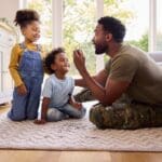 A trucking dad sitting on the floor with a puzzle piece in his hand as he helps his two daughters work on a puzzle together
